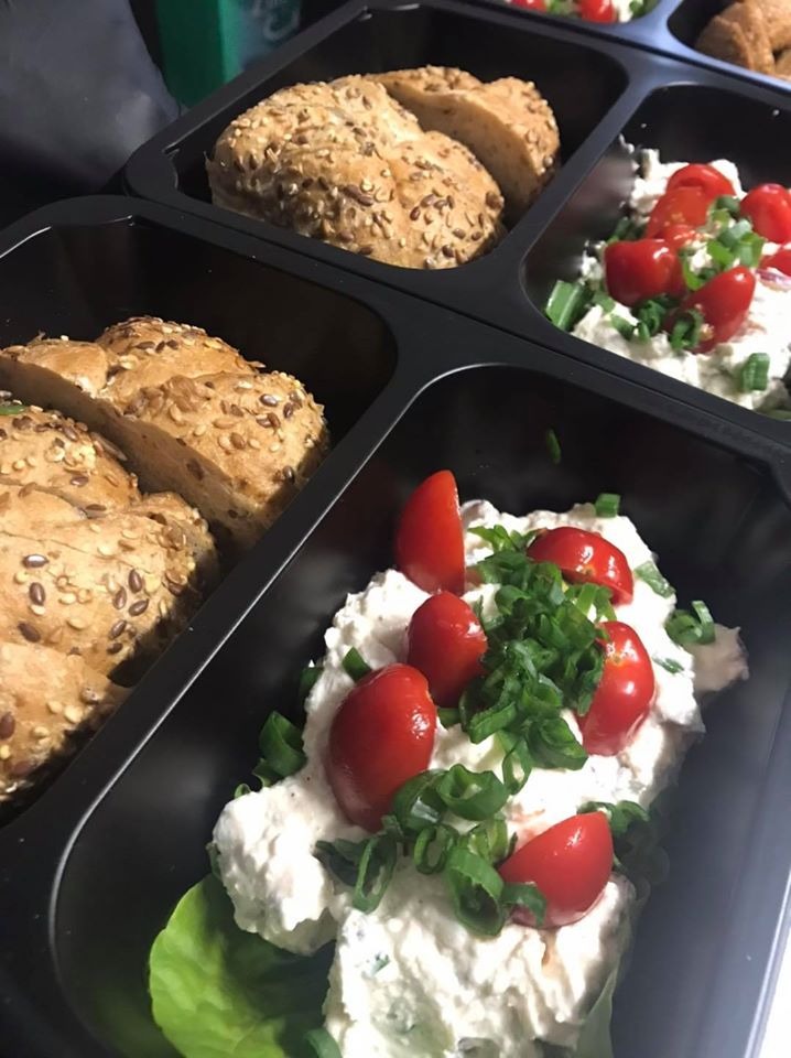 Porcje w czarnych pojemnikach: pieczywo pełnoziarniste i pasta twarogowa z pomidorkami koktajlowymi i szczypiorkiem na sałacie.