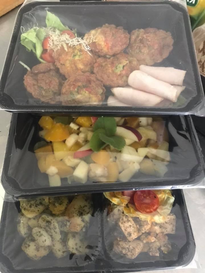 Zestaw trzech czarnych pudełek z przezroczystym wieczkiem, zawierających różne gotowe posiłki: kotleciki z warzywami i sałatką, sałatka owocowa z miętą i danie z ziemniaków i tofu.