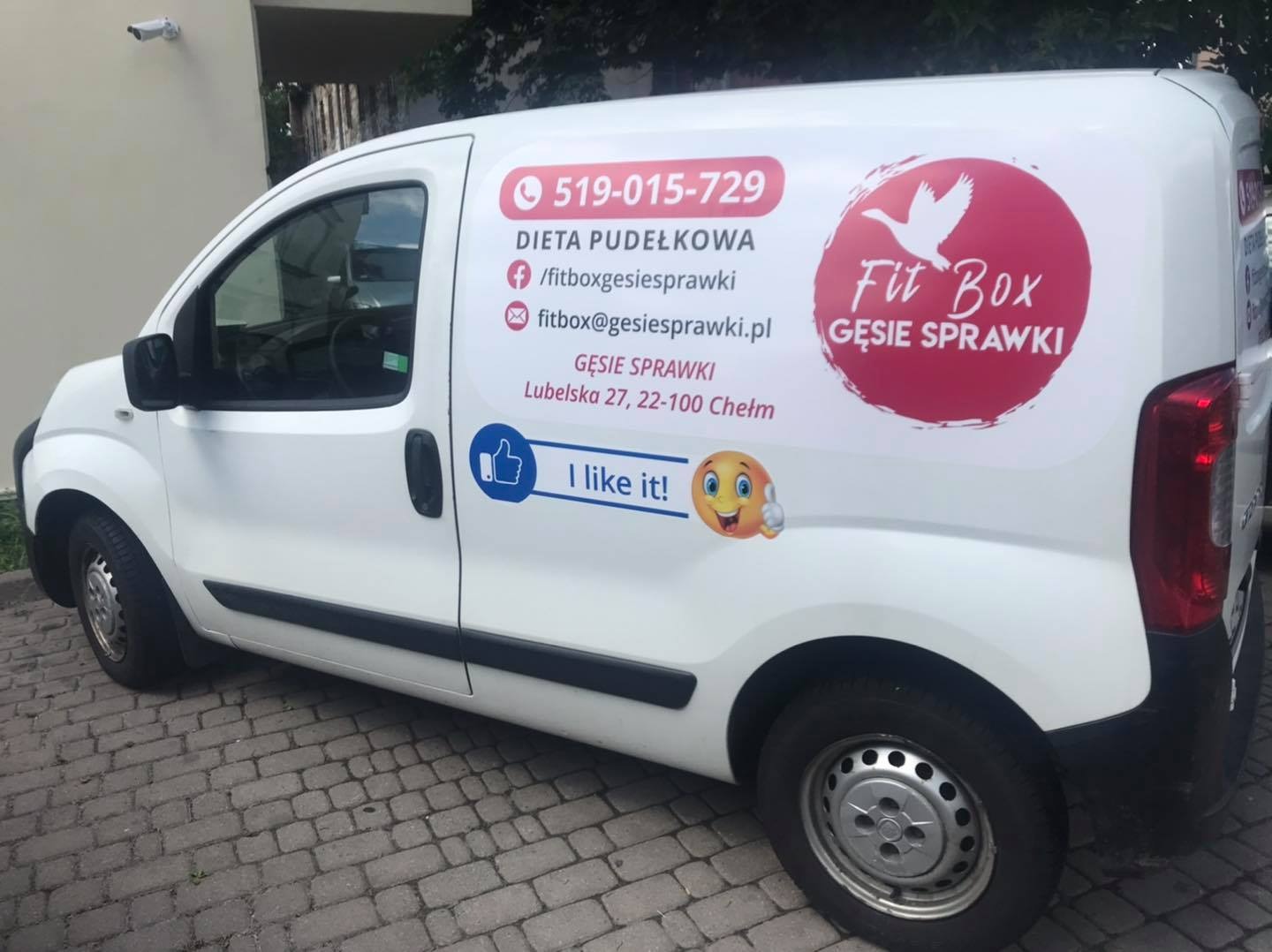Biały samochód dostawczy z reklamą diety pudełkowej, widoczny numer telefonu, adres e-mail i logo firmy Fit Box Gęsie Sprawki.