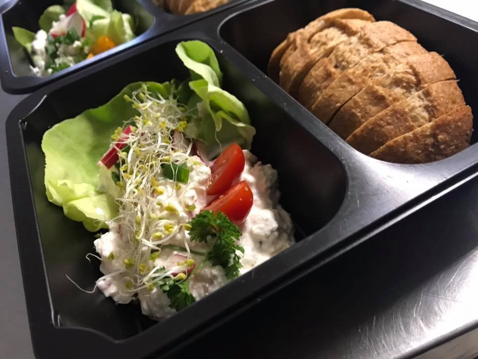 Pudełko z gotowym posiłkiem z Chełma: twarożek ze świeżymi warzywami i kiełkami na liściu sałaty, obok pieczywo razowe.