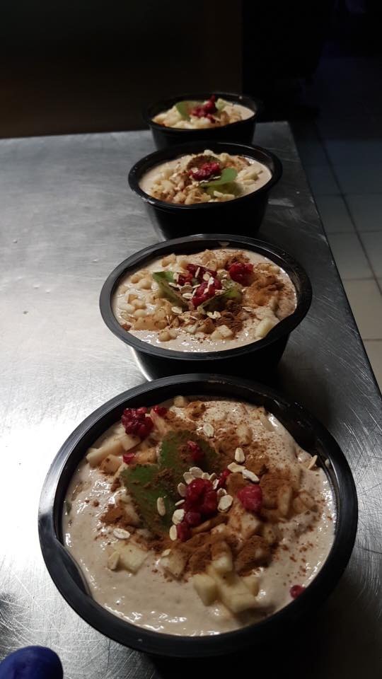 Cztery porcje kremowego deseru w czarnych miseczkach, udekorowane kawałkami jabłek, malinami, płatkami owsianymi, miętą i posypane cynamonem, ustawione w rzędzie na metalowym blacie.