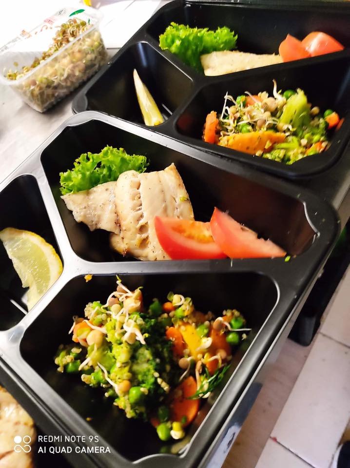 Kompozycja dań w czarnych, wielodzielnych pojemnikach: grillowana ryba z cytryną i pomidorem, sałatka warzywna z kiełkami, zielona sałata z tofu.