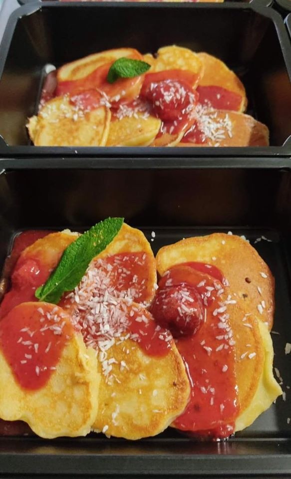 Dwa czarne pojemniki z pankejkami polanymi sosem truskawkowym, posypane wiórkami kokosowymi i udekorowane listkiem mięty.