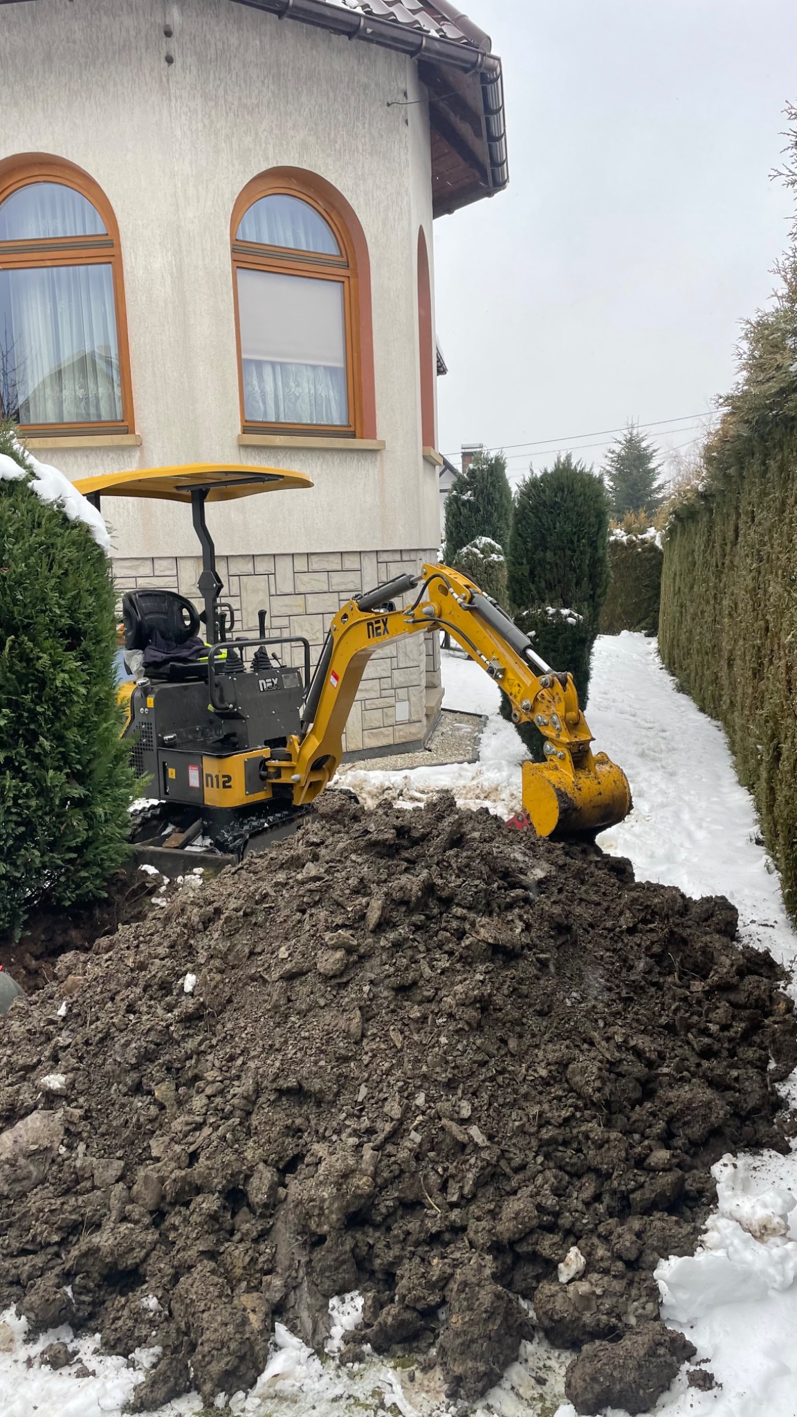 Żółta mini koparka NEX N12 pracuje na zasypanym śniegiem terenie, obok domu z oknami w kształcie łuku i żywopłotu.