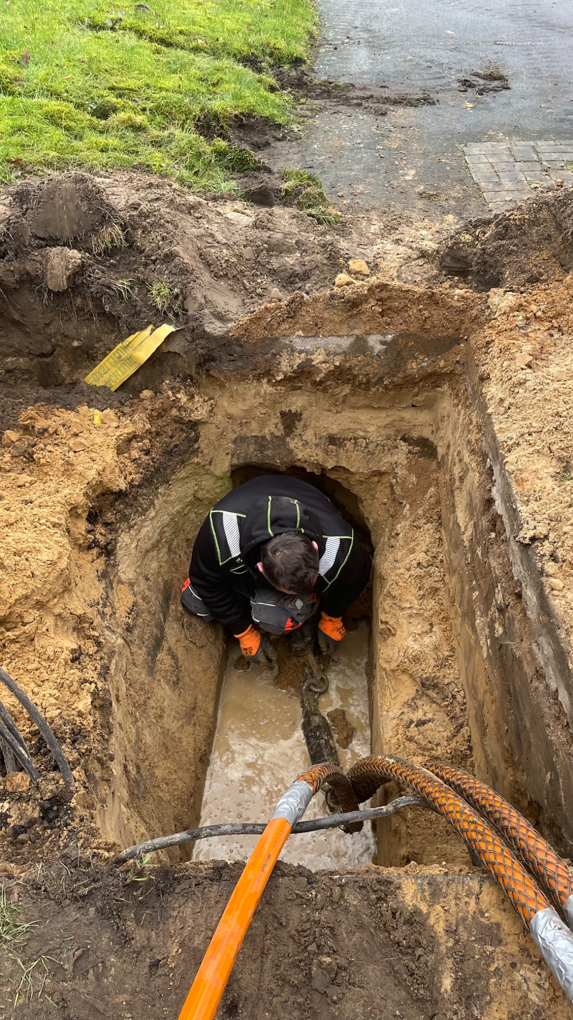 Pracownik w pomarańczowych rękawicach w głębokim wykopie wypełnionym wodą, instaluje kable telekomunikacyjne z pomarańczową osłoną, widoczne narzędzia i żółta taśma ostrzegawcza przy krawędzi wykopu.