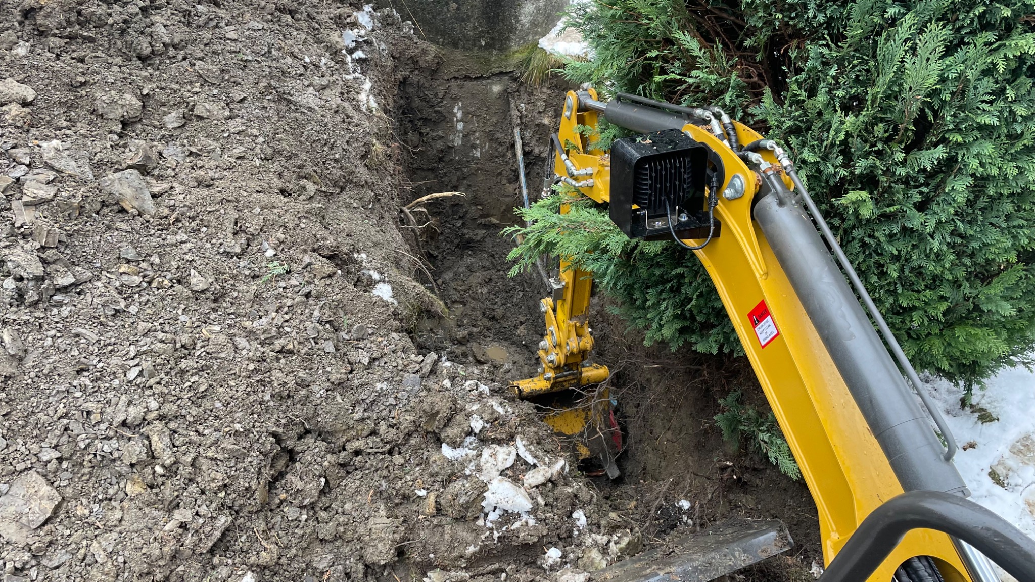 Żółta mini koparka wykopuje wąski rów w ziemi obok zielonego krzewu, widoczne fragmenty śniegu i błoto.