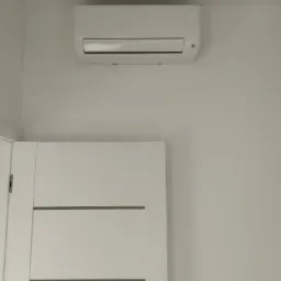 Biała jednostka klimatyzacyjna zamontowana na ścianie nad białą, nowoczesną, wewnętrzną klatką drzwiową z poziomymi wgłębieniami.