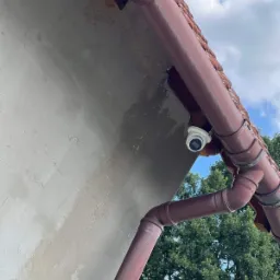 Kamera monitoringu zamontowana pod okapem budynku z brązową rynną, widok z dołu na tle błękitnego nieba i zielonych drzew.