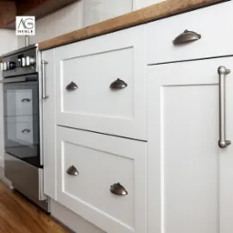 Białe szafki kuchenne z drewnianym blatem, metalowymi uchwytami typu muszelka i pionowym uchwytem rurowym, sąsiadujące ze stalowym piekarnikiem.