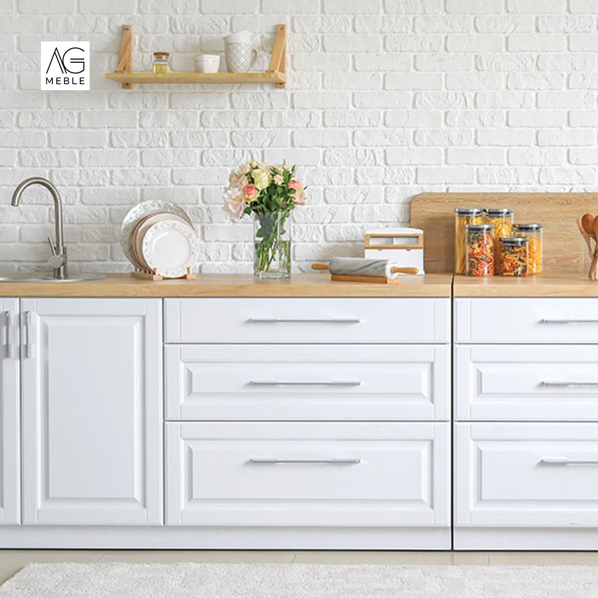 Białe, klasyczne szafki kuchenne z drewnianym blatem, na którym stoją akcesoria kuchenne i wazon z kwiatami, na tle ściany z białej cegły.