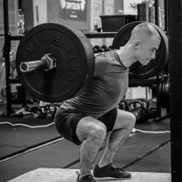 Radosław Rudnicki - Trener Personalny - Czarno-białe zdjęcie mężczyzny wykonującego przysiad ze sztangą w siłowni, widoczne kettlebells w tle.