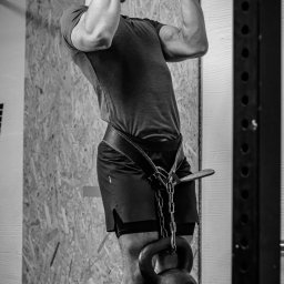 Radosław Rudnicki - Trener Personalny - Mężczyzna wykonuje podciąganie na drążku z obciążeniem w postaci kettlebell zawieszonego na pasie, widoczne napięte mięśnie ramion i pleców, czarno-biała fotografia.