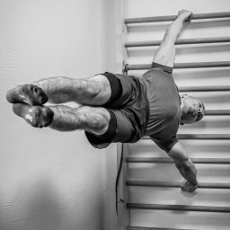 Radosław Rudnicki - Trener Personalny - Mężczyzna w spodenkach i skarpetach wspinaczkowych wykonuje ćwiczenie na drabinkach gimnastycznych, trzymając się jedną ręką i jedną nogą, w czarno-białej tonacji.