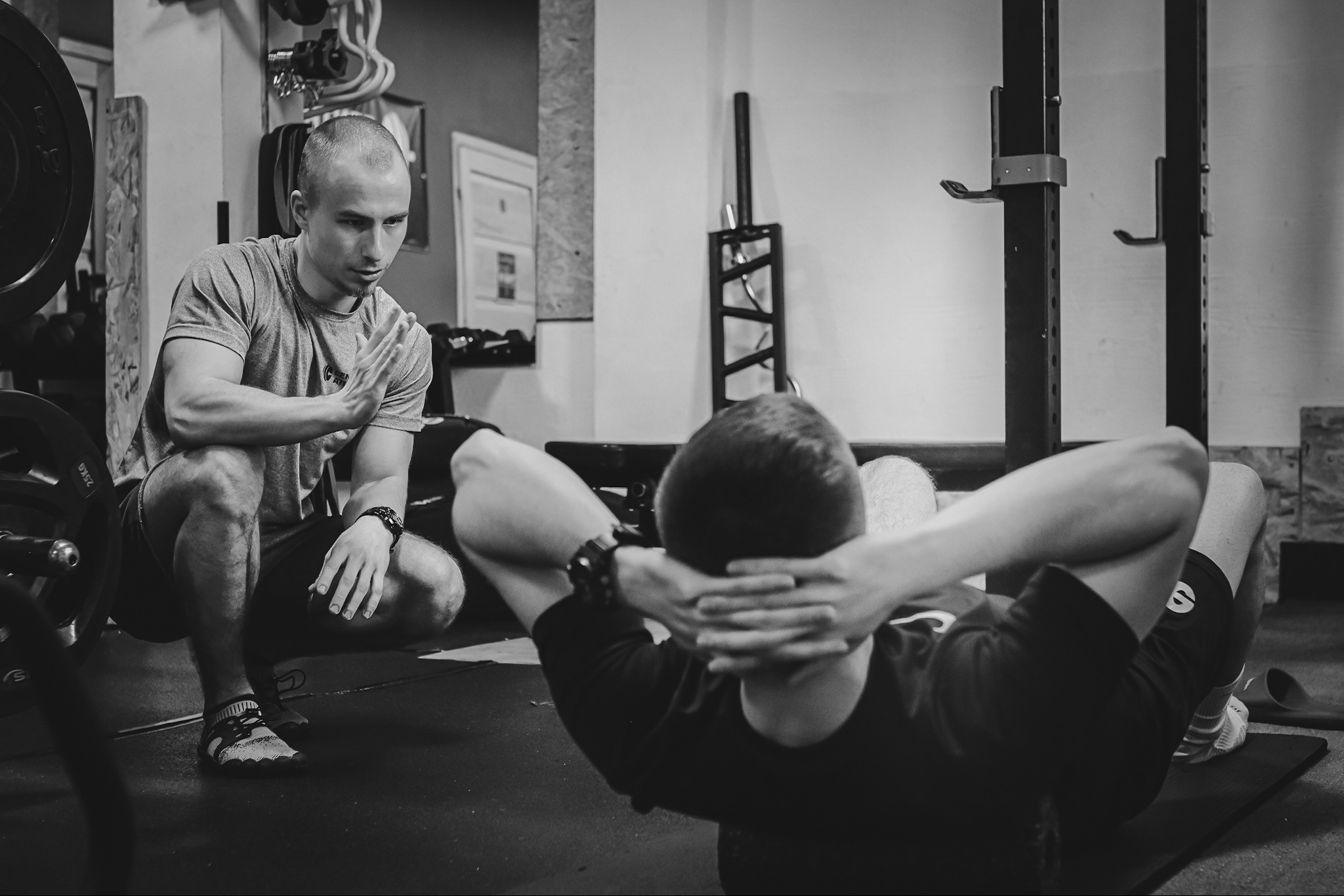 Czarno-białe zdjęcie: Trener personalny obserwuje mężczyznę wykonującego ćwiczenia na mięśnie brzucha na macie w siłowni, widoczne sztangi i sprzęt do ćwiczeń.