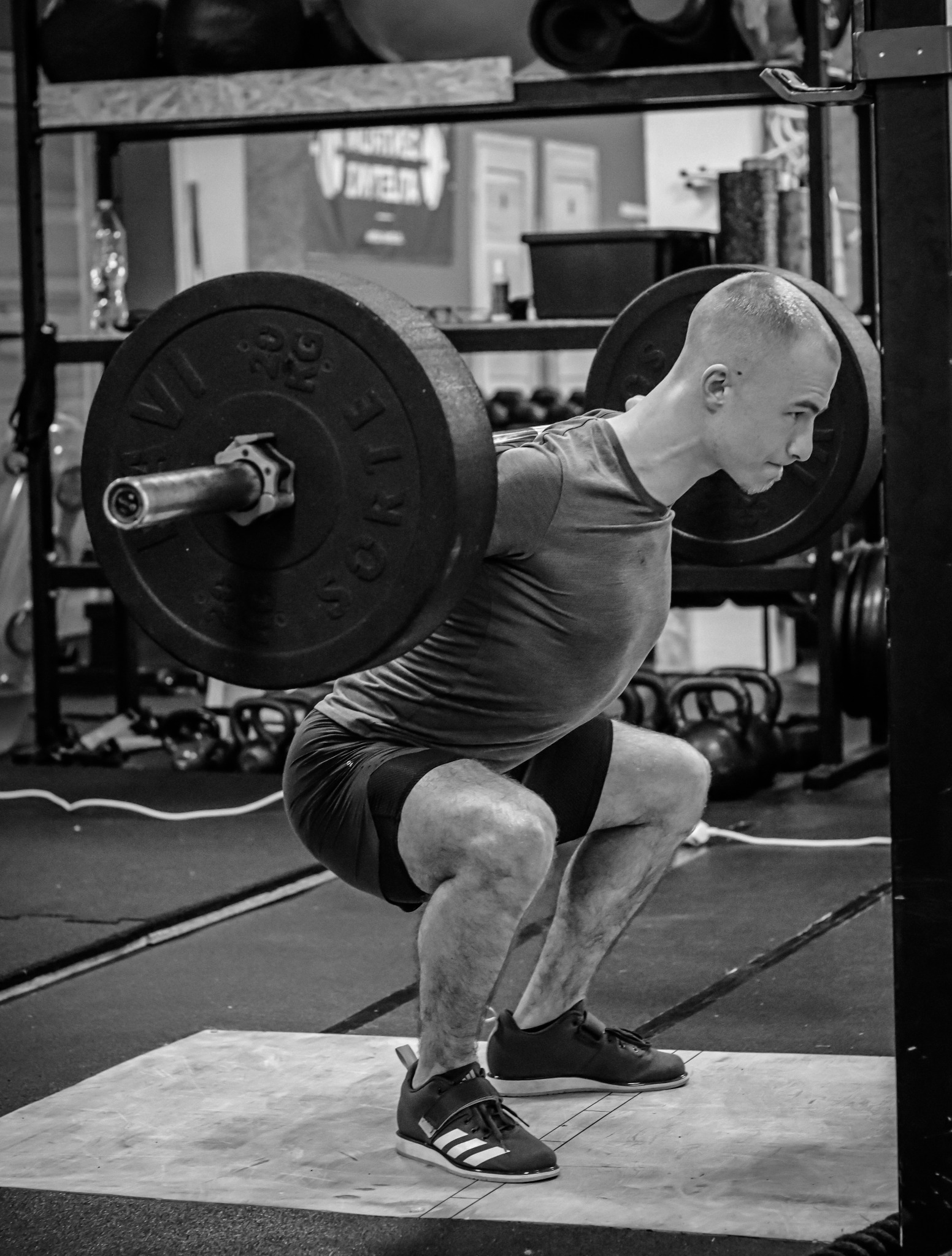 Czarno-białe zdjęcie mężczyzny wykonującego przysiad ze sztangą w siłowni, widoczne kettlebells w tle.