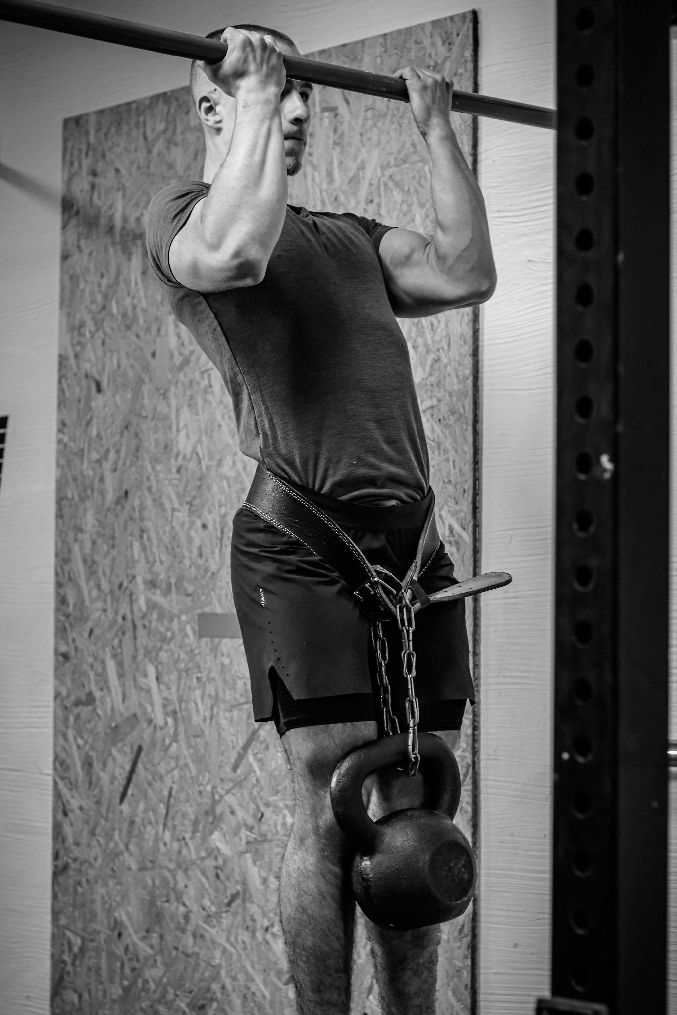 Mężczyzna wykonuje podciąganie na drążku z obciążeniem w postaci kettlebell zawieszonego na pasie, widoczne napięte mięśnie ramion i pleców, czarno-biała fotografia.