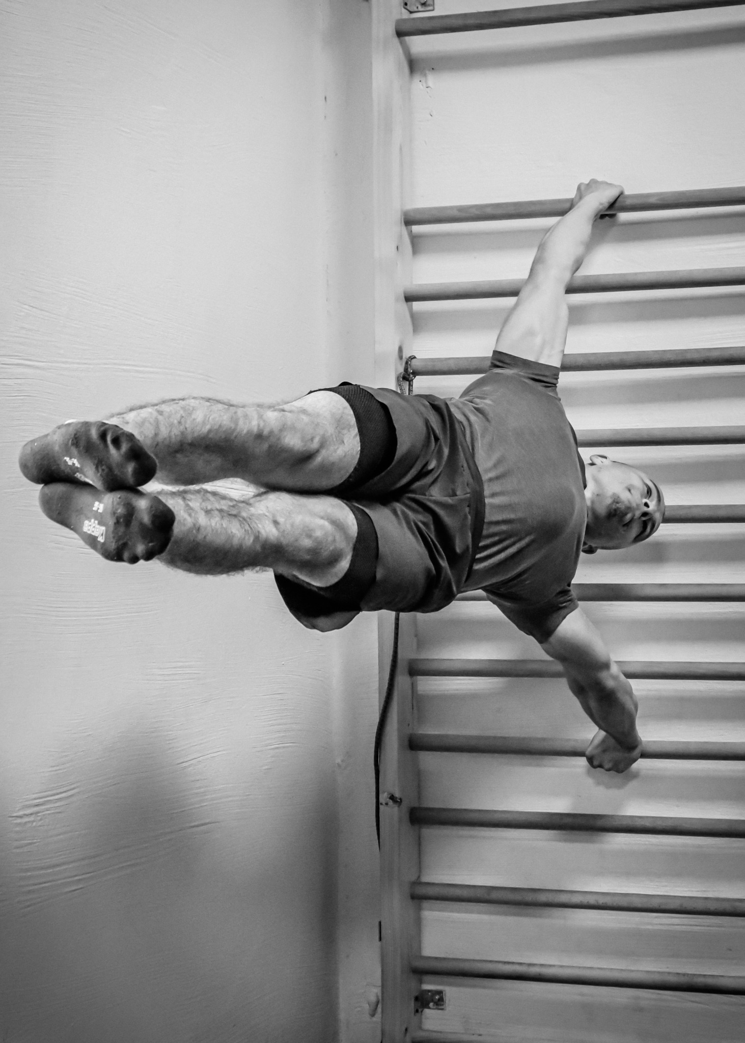 Mężczyzna w spodenkach i skarpetach wspinaczkowych wykonuje ćwiczenie na drabinkach gimnastycznych, trzymając się jedną ręką i jedną nogą, w czarno-białej tonacji.