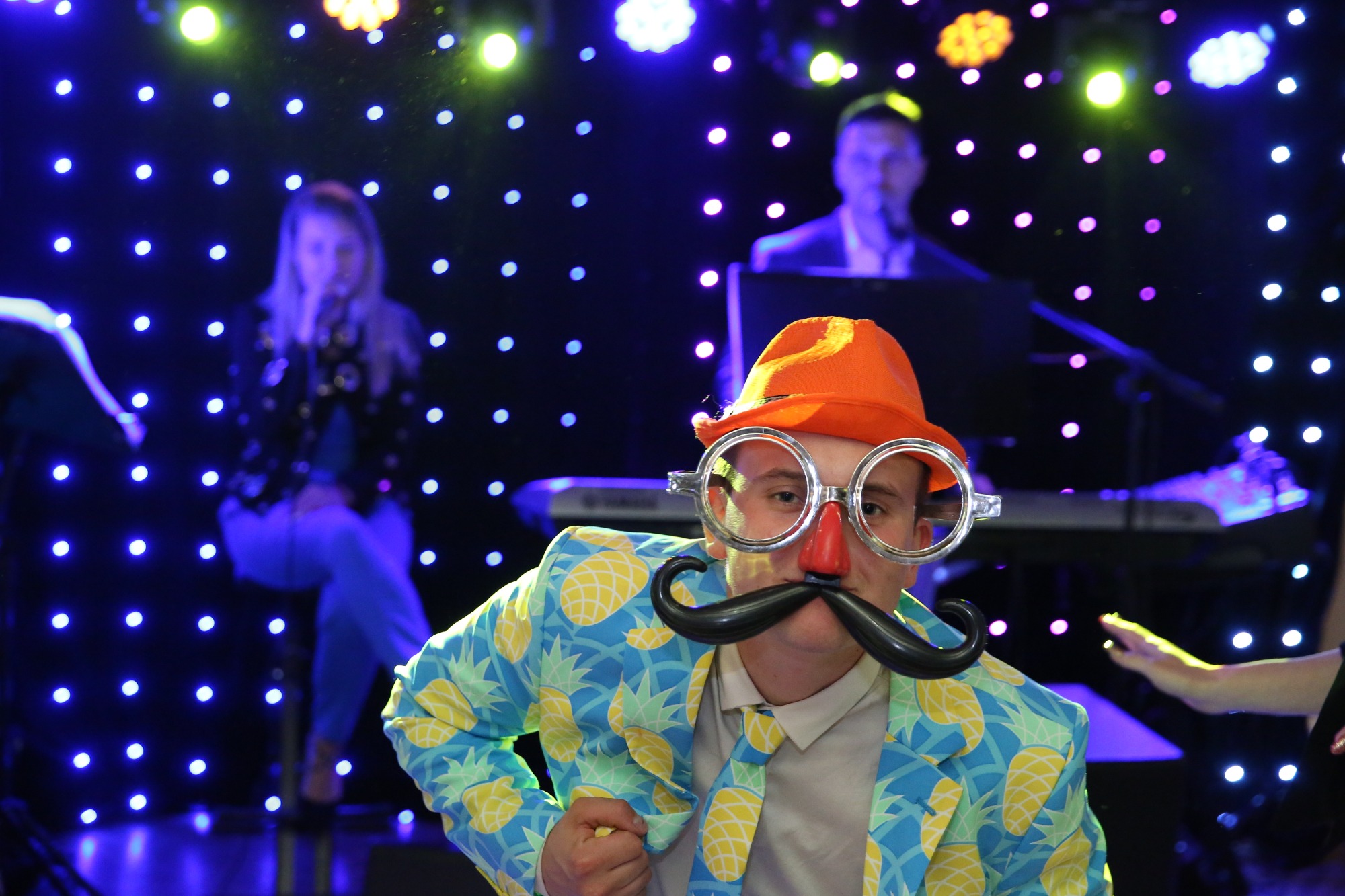 Osoba w przebraniu z pomarańczowym kapeluszem, okularami z nosem klauna i sztucznym wąsem, w marynarce w ananasy, na tle dekoracji świetlnych i innych osób, prawdopodobnie podczas imprezy.