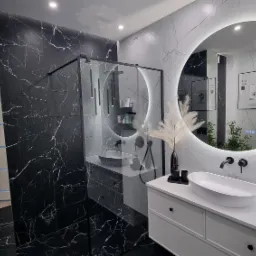 Nowoczesna łazienka z czarno-białymi płytkami imitującymi marmur, okrągłym lustrem z podświetleniem LED, umywalką nablatową i kabiną prysznicową typu walk-in z czarnymi profilami.
