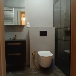 Nowoczesna łazienka we Wrocławiu z podwieszaną toaletą z bidetką w kolorze złotym, czarną szafką pod umywalką i kabiną prysznicową z czarnymi ramami.