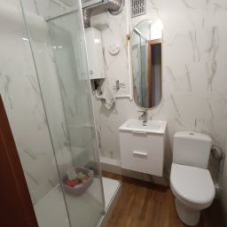 Remonty i Wykończenia Wnętrz Wrocław - Nowoczesna łazienka z kabiną prysznicową, umywalką z szafką, toaletą i lustrem w złotej ramie. Ściany wykończone marmurową płytką, podłoga drewnopodobna.