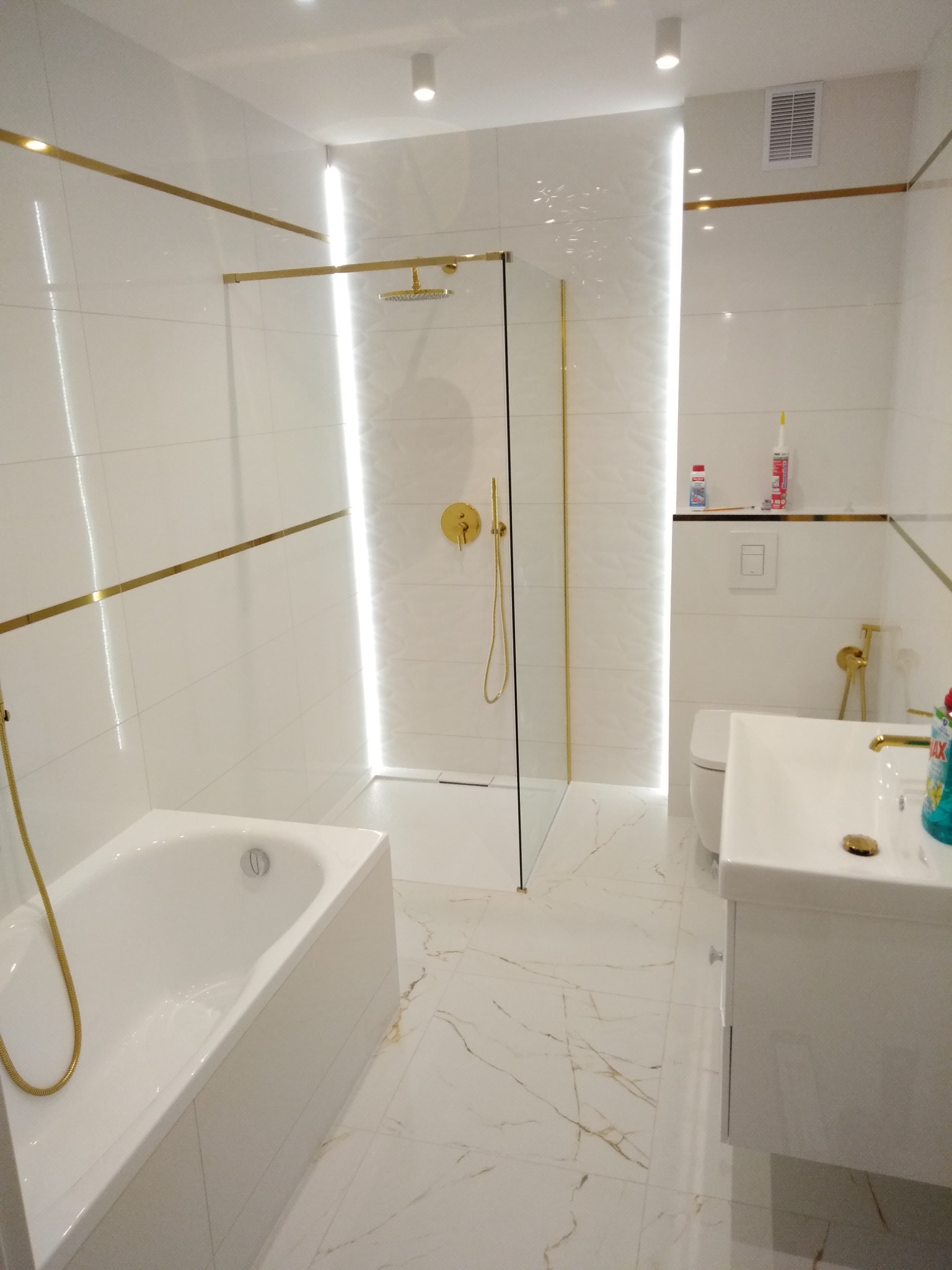 Nowoczesna łazienka z białymi płytkami, złotymi akcentami, kabiną prysznicową typu walk-in i marmurową podłogą.