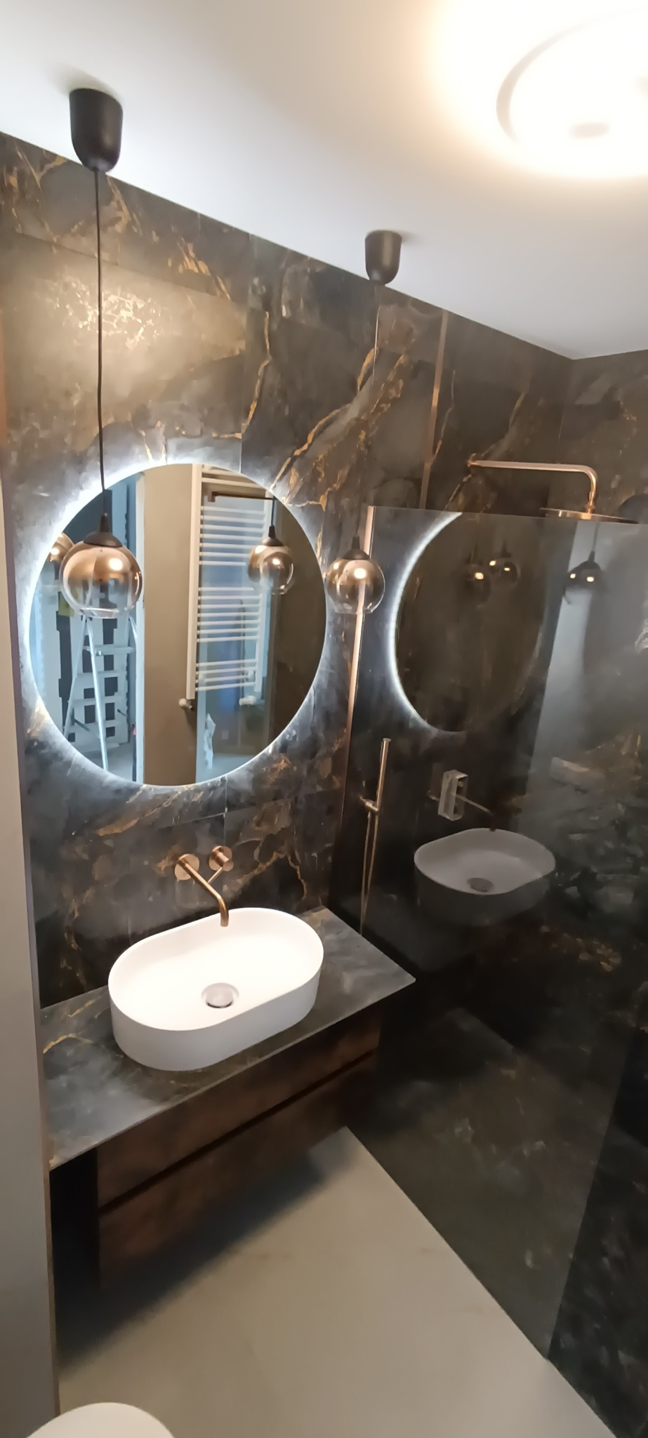 Elegancka łazienka z czarnymi marmurowymi ścianami, okrągłym lustrem z podświetleniem LED, białą umywalką nablatową i złotą armaturą; w lustrze odbija się drabina.