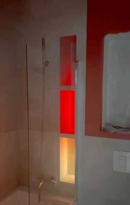 Nowoczesna łazienka z kabiną prysznicową i wnękami podświetlonymi na czerwono i pomarańczowo. Minimalistyczny design z akcentem kolorystycznym.