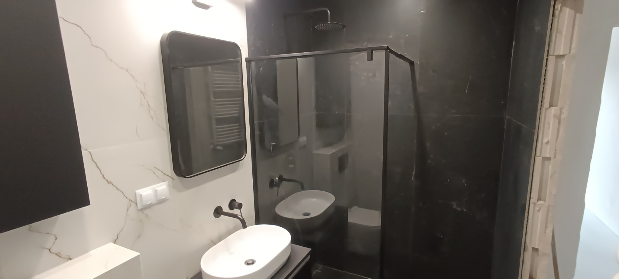 Nowoczesna łazienka z czarną armaturą, owalną umywalką nablatową i kabiną prysznicową typu walk-in. Marmurowe płytki na ścianach dodają elegancji.