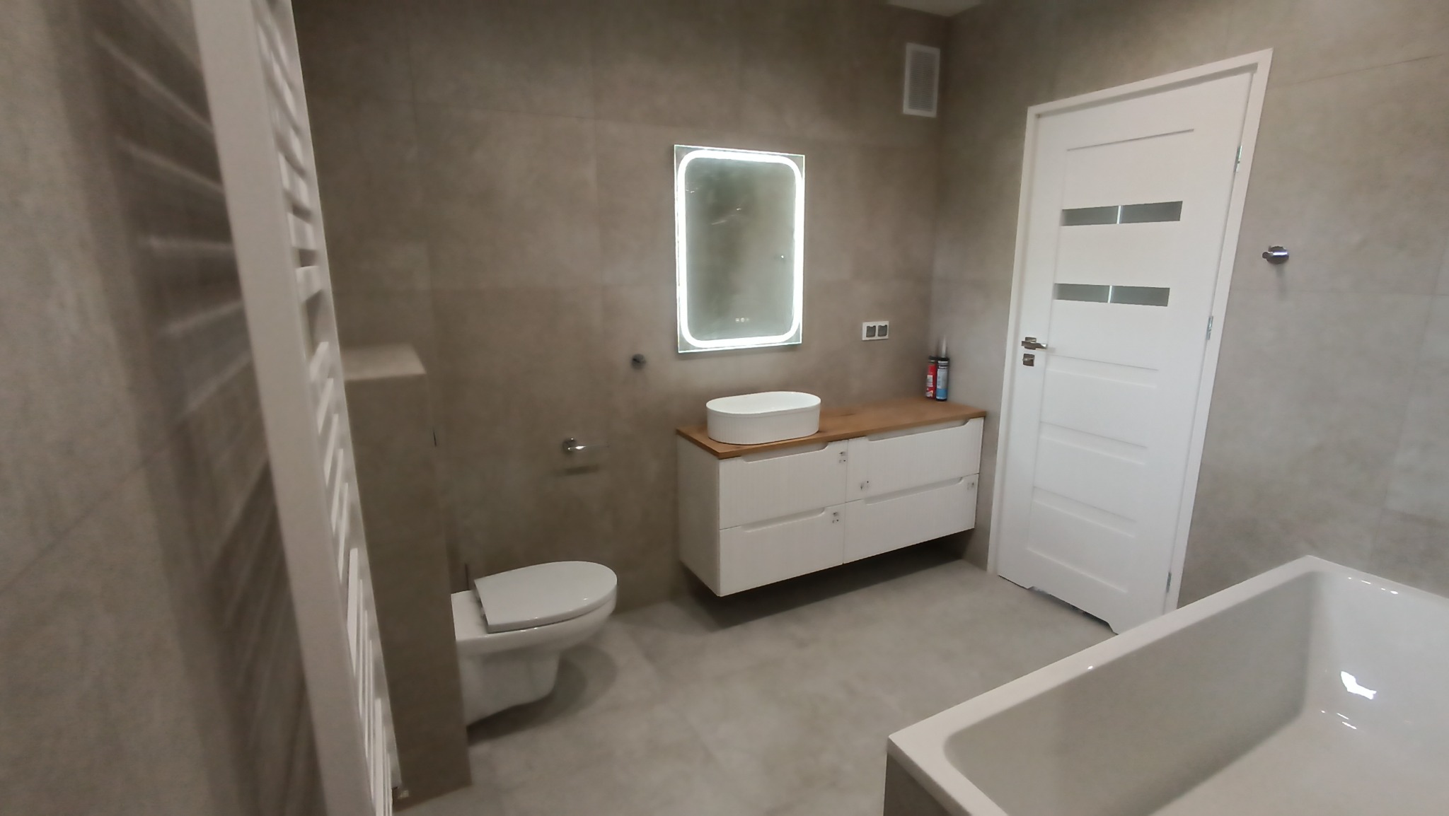 Nowoczesna łazienka z beżowymi płytkami, białymi meblami i podświetlanym lustrem. Widoczna toaleta, umywalka nablatowa i fragment wanny. Minimalistyczny design.