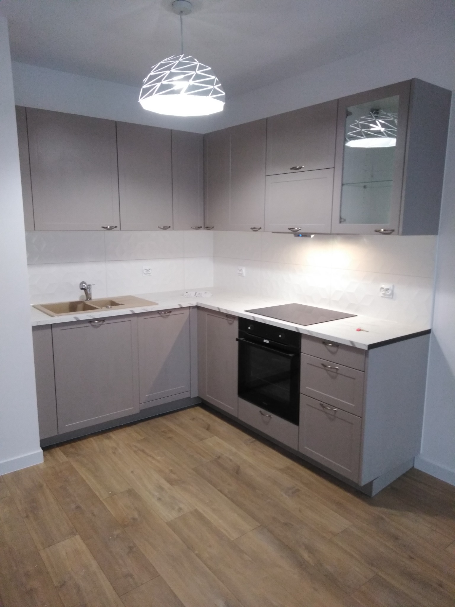 Nowoczesna kuchnia w odcieniach szarości z drewnianą podłogą, oświetlona lampą wiszącą o geometrycznym wzorze, z widocznym zlewem, płytą indukcyjną i piekarnikiem.
