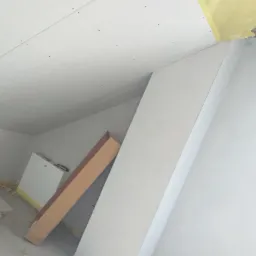 Skośne poddasze z zamontowaną płytą gipsowo-kartonową, widoczne łączenia płyt, niezabezpieczone narożniki, kartonowe opakowania na podłodze, żółta taśma malarska wzdłuż krawędzi.