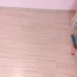 Jasne panele podłogowe z widocznym opakowaniem Kronopol Room, szczotką do zamiatania i fragmentem drabiny malarskiej przy różowej ścianie.