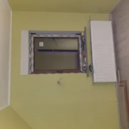 Montaż okna PCV z drewnopodobną ramą w ścianie o limonkowym odcieniu, widoczne wystające przewody elektryczne i grzejnik panelowy obok okna.