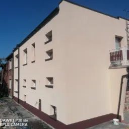 Elewacja budynku po remoncie w Pińczowie, z nową, jasną elewacją i ciemnobrązowym cokołem, widoczna drabina na balkonie i satelita.