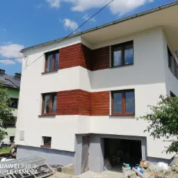 Dwukondygnacyjny dom z białą elewacją i drewnianymi panelami dekoracyjnymi, widoczne materiały budowlane przed garażem, sugerujące prace wykończeniowe lub instalacyjne