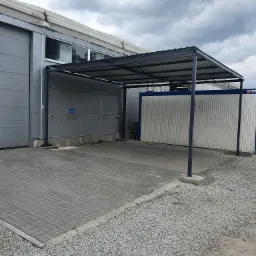 Zadaszenie metalowe w kolorze grafitowym, konstrukcja wsparta na czterech słupach, przylegająca do budynku magazynowego i kontenerów, wyłożona kostką brukową i żwirem.