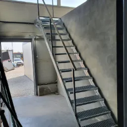 Metalowe schody kratowe z czarnymi poręczami prowadzące na antresolę w hali przemysłowej, widok z dołu na tle betonowej ściany i okna z widokiem na plac.