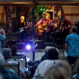 Zespół muzyczny grający bluesa na scenie podczas koncertu plenerowego, widownia siedząca przy stolikach, oświetlenie sceniczne i dekoracje.