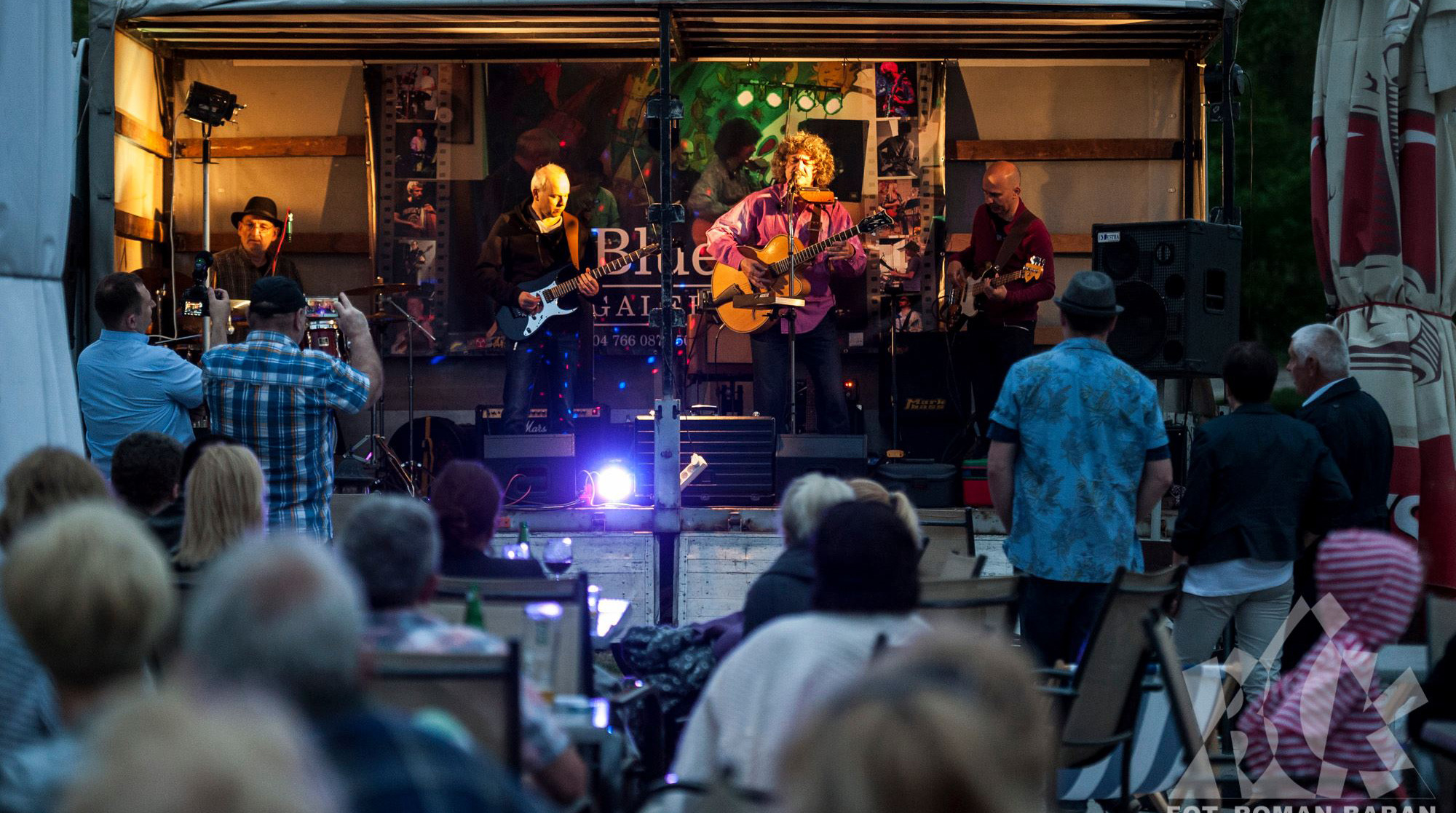 Zespół muzyczny grający bluesa na scenie podczas koncertu plenerowego, widownia siedząca przy stolikach, oświetlenie sceniczne i dekoracje.