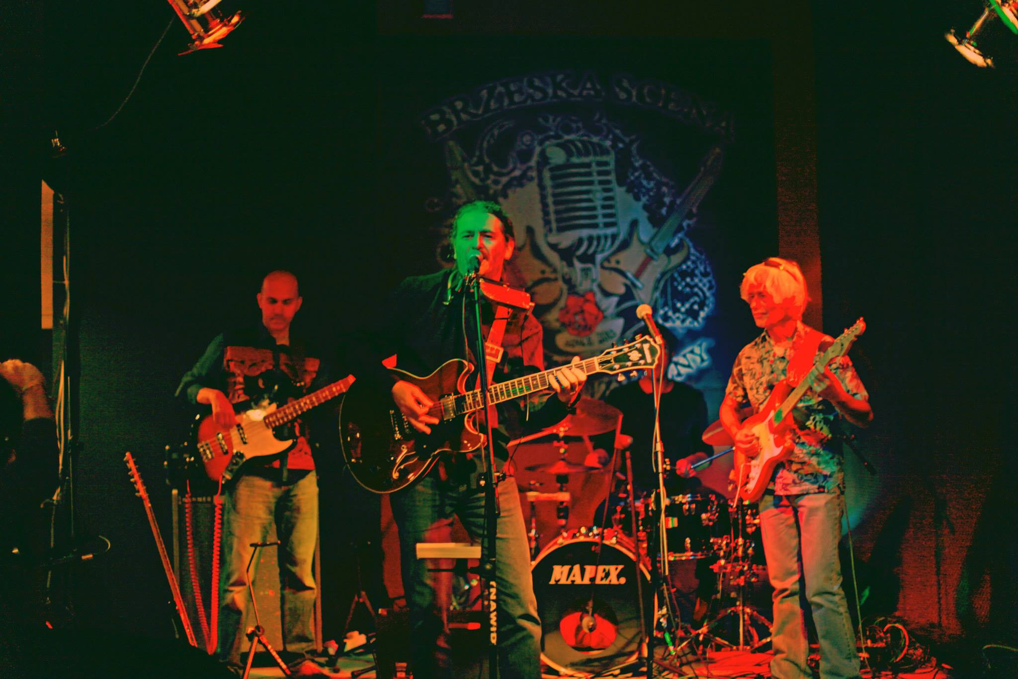 Zespół muzyczny gra koncert na scenie, wokalista śpiewa do mikrofonu i gra na gitarze, w tle perkusja Mapex i logo 'Brzeska Scena'