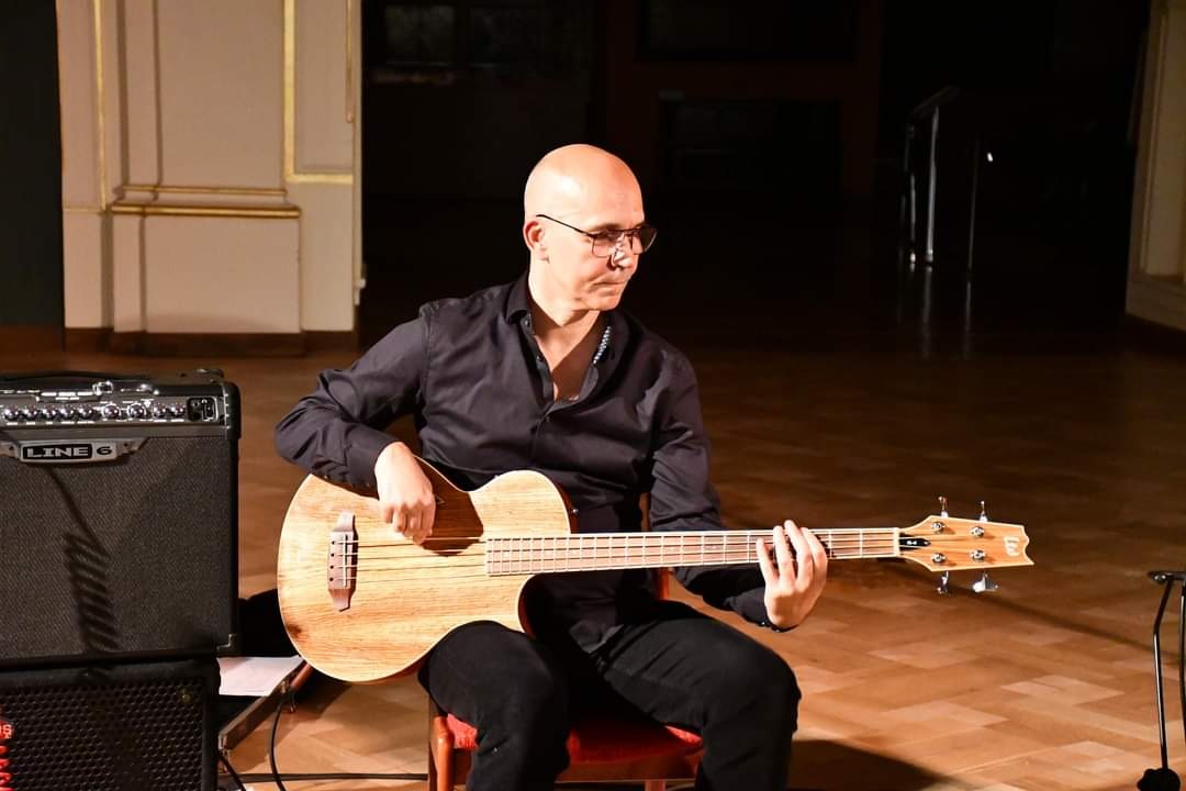 Mężczyzna w okularach gra na gitarze basowej akustycznej, siedząc na krześle. W tle wzmacniacz Line 6 i parkiet. Scena oświetlona ciepłym światłem.