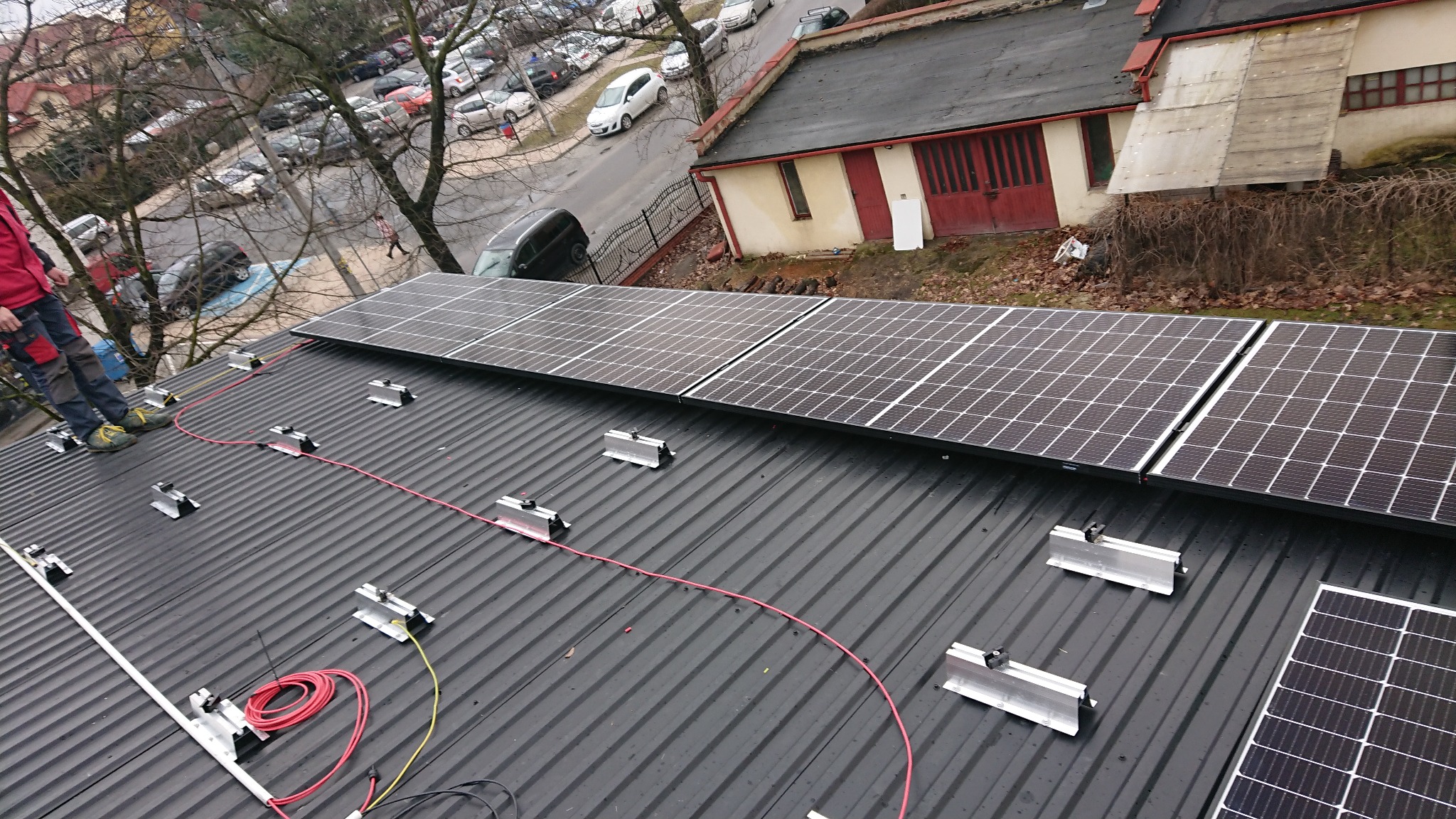 Montaż paneli słonecznych na dachu z blachy trapezowej, widoczne mocowania, kable i pracownik w czerwonej kurtce.