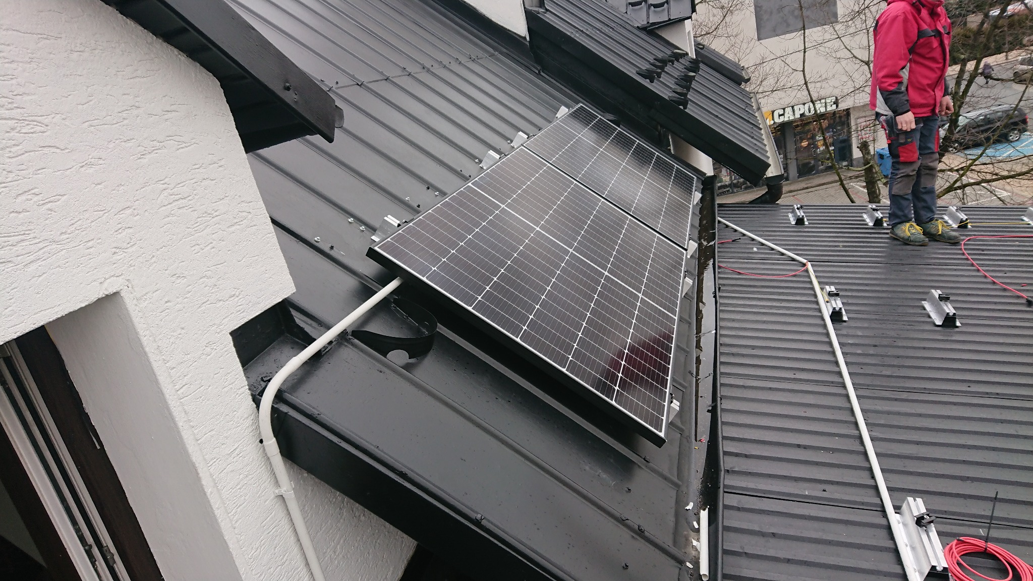 Montaż paneli fotowoltaicznych na czarnym blaszanym dachu, widoczne elementy mocujące, biała rura osłonowa i częściowo sylwetka instalatora w czerwonej kurtce.