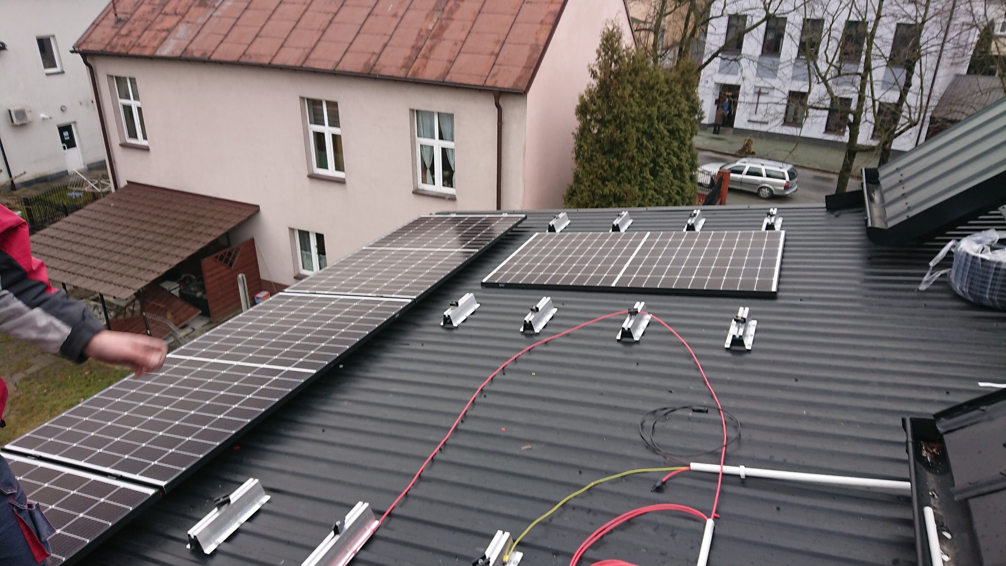 Montaż paneli słonecznych na dachu z blachy trapezowej, widoczne kable i elementy mocujące, w tle budynki mieszkalne i drzewa.