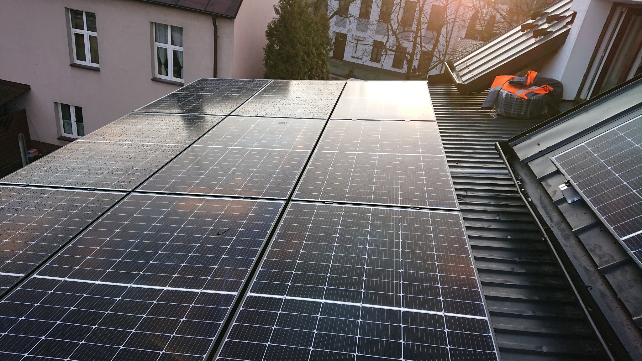 Panele słoneczne na dachu budynku mieszkalnego, niektóre z kroplami deszczu, w tle budynki i drzewa, w rogu dachu leży pomarańczowa kamizelka odblaskowa.