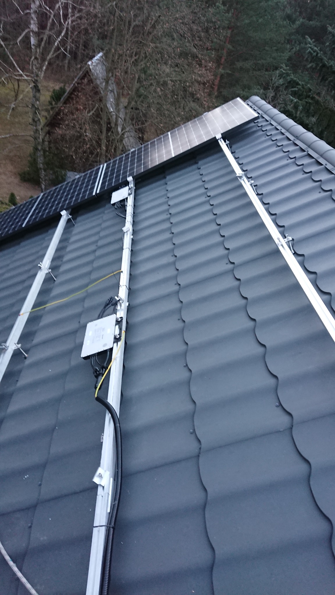 Panele słoneczne zamontowane na ciemnoszarym dachu z falistej blachy, widok z góry, w tle drzewa bez liści.
