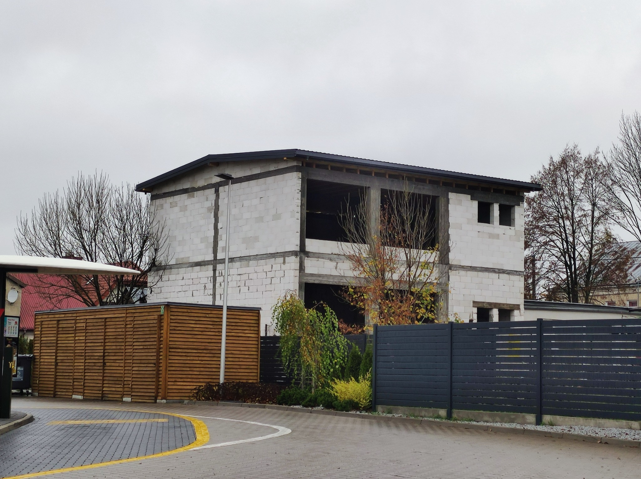 Dwukondygnacyjny budynek w stanie surowym otwartym z białych bloczków, widoczne okna bez ram, czarny dach, szara kostka brukowa i drewniane ogrodzenie w tle.