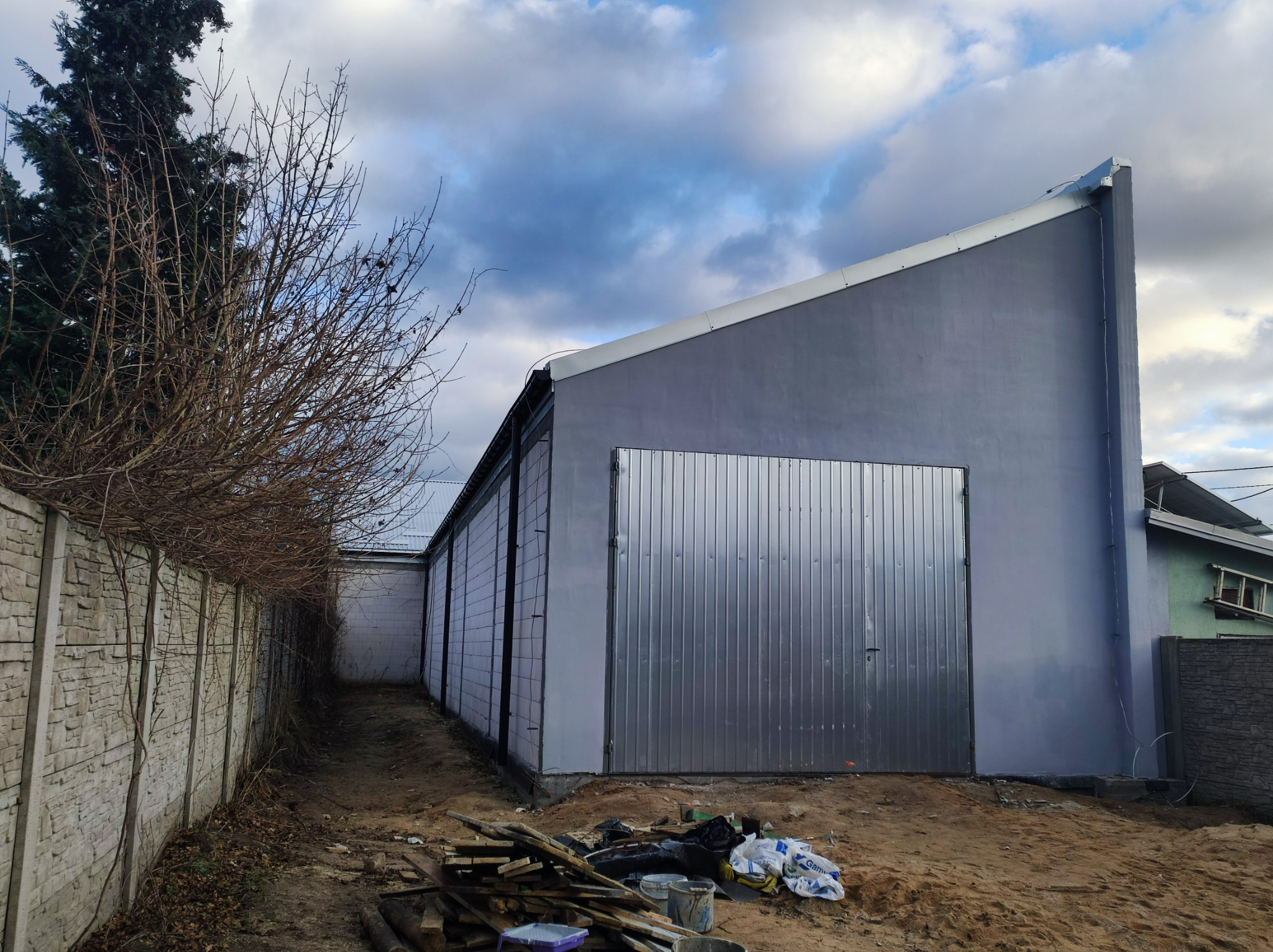 Szary budynek gospodarczy z metalową bramą garażową, widoczny z boku, z przygotowanym terenem i materiałami budowlanymi przed nim.