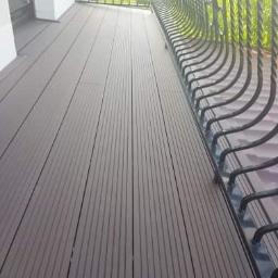 DachNel - Nowoczesny taras z desek kompozytowych w odcieniach brązu, widok z góry na balustradę z metalowymi zdobieniami, jasne tło ściany budynku.